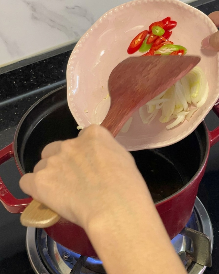 Geschnittene rote und grüne Chilischoten sowie Kräuter werden mit einem Holzspatel von einem kleinen rosa Teller in einen geölten roten Topf geschoben.