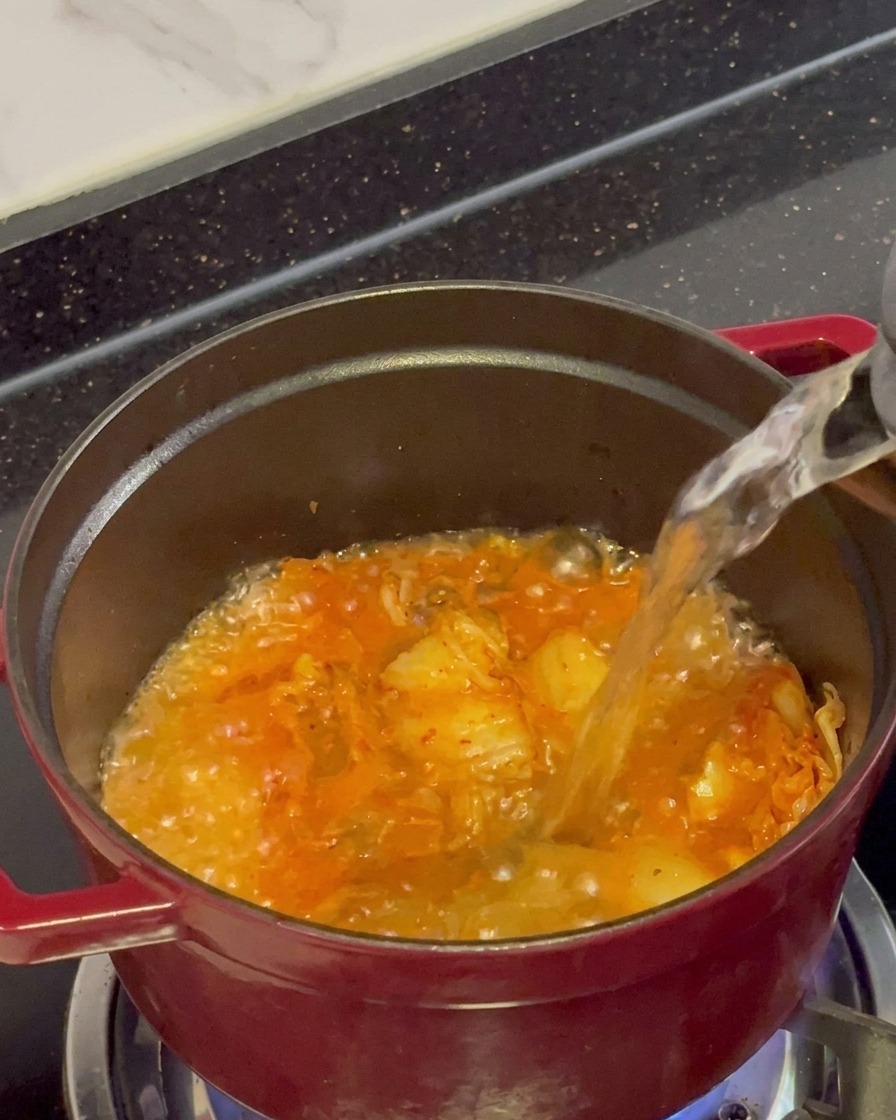 Klares, kochendes Wasser wird aus einem Wasserkocher in einen roten Topf mit brodelndem, angebratenem Kimchi gegossen.