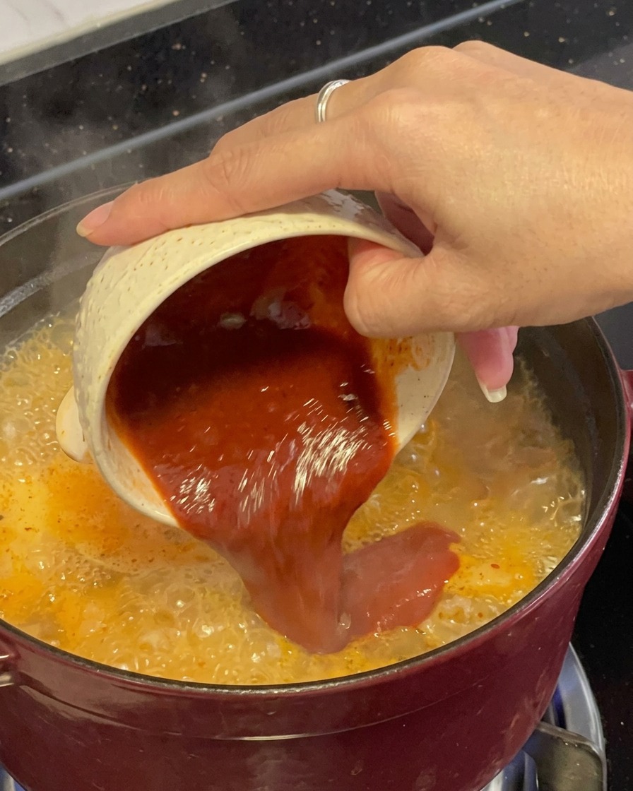 Eine dicke, dunkelrote Gewürzmischung wird aus einer kleinen gemusterten Schale direkt in den kochenden roten Meeresfrüchte-Eintopf gegossen.