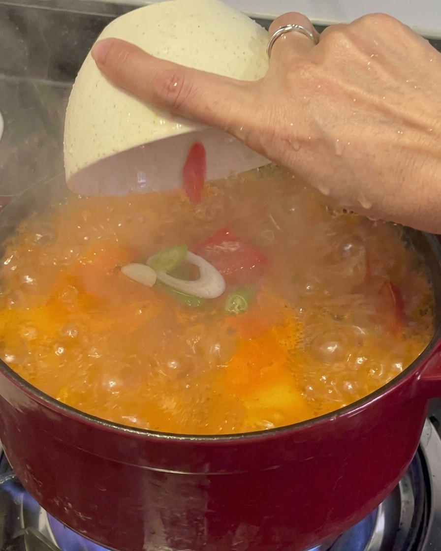 Frisch geschnittene grüne Frühlingszwiebeln und rote Chilischoten werden aus einer weißen Schale über den dampfenden, kochenden Kimchi-Eintopf gestreut.