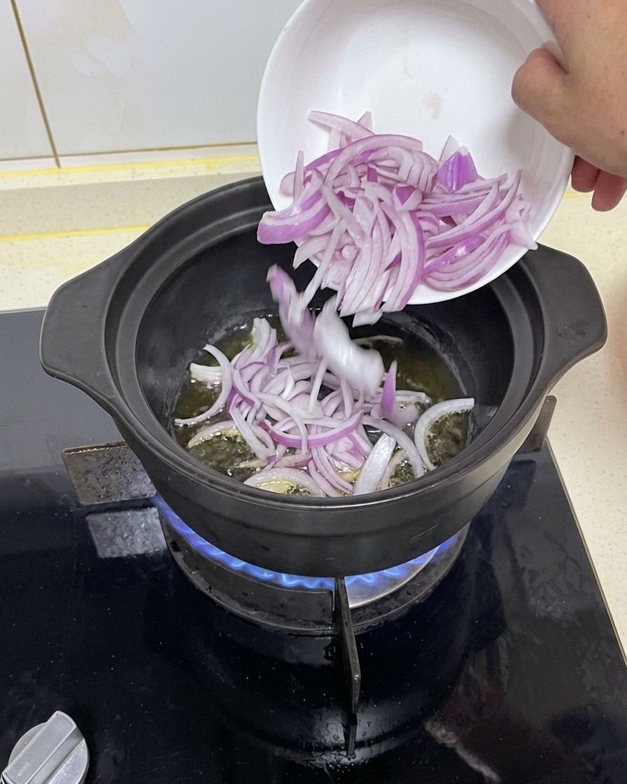Rote Zwiebelspalten werden aus einer kleinen weißen Schüssel in einen heißen, schwarzen Kochtopf mit Öl über einer blauen Gasflamme gegeben.