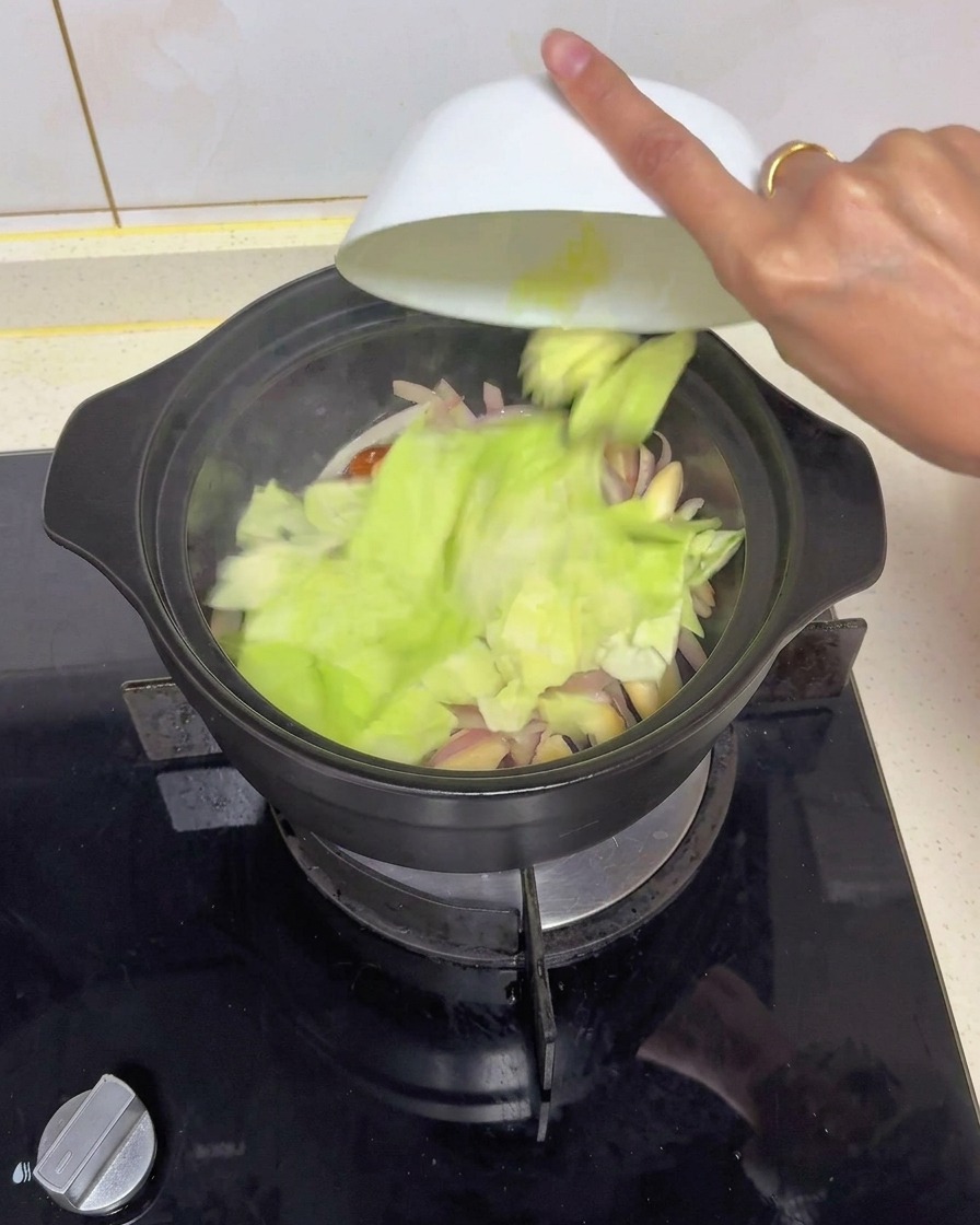 Grob geschnittene Stücke von frischem Weißkohl werden aus einer weißen Schüssel in einen schwarzen Topf mit angebratenen Zwiebeln und Pilzen geschüttet.
