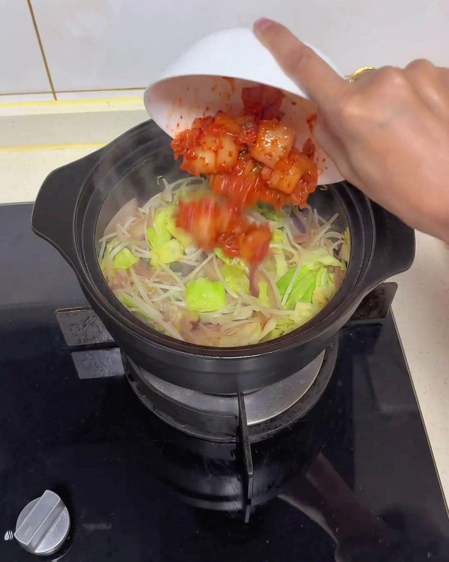 Eine Hand kippt eine weiße Schüssel, um leuchtend rotes, gehacktes Kimchi in einen schwarzen Tontopf voller glasiger Zwiebeln, Weißkohl und Sojasprossen zu geben.