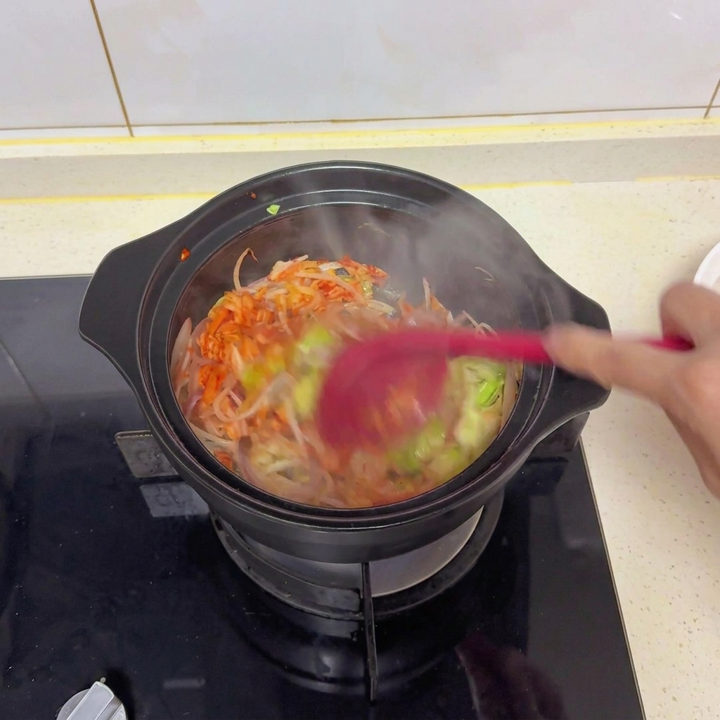 Gemischtes Gemüse und Kimchi werden in einem schwarzen Topf mit einem roten Pfannenwender umgerührt, um Salz, Sojasauce und Gewürze zu verteilen.