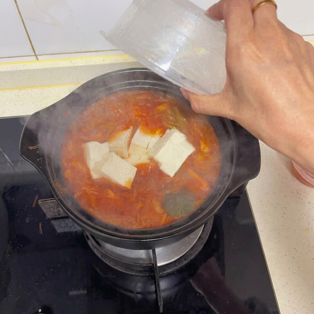 Eine Hand gibt vorsichtig große Würfel von weißem Tofu aus einer klaren Plastikverpackung in einen kochenden Topf mit roter Kimchi-Suppe.