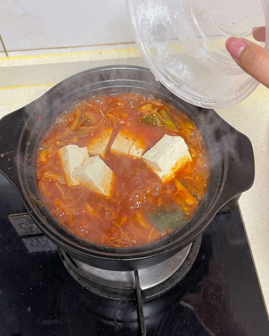 Weiße, rechteckige Tofustücke liegen in einem kochenden Topf mit tiefroter Kimchi-Suppe, umgeben von Gemüse und Seetang.