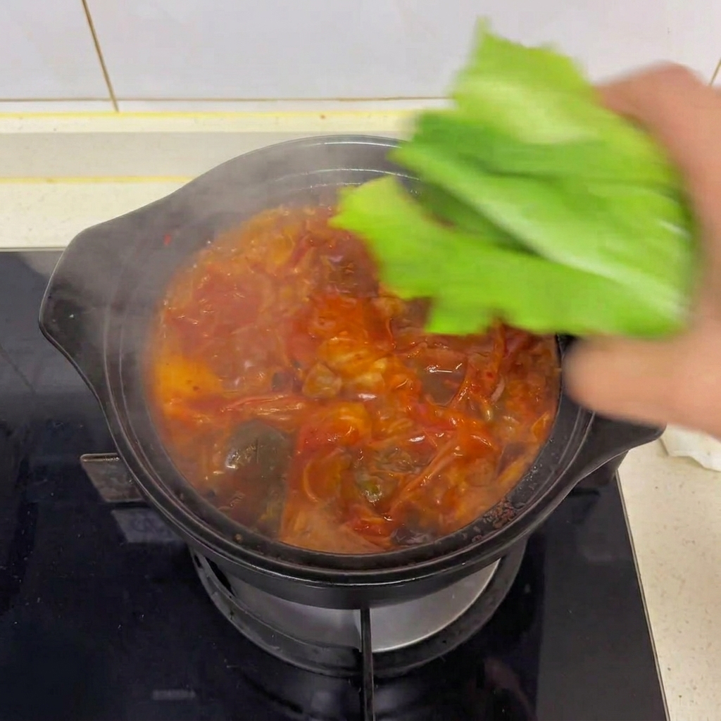 Eine Hand gibt frische grüne Römersalatblätter in einen dampfend heißen Topf mit Kimchi-Tofu-Suppe.