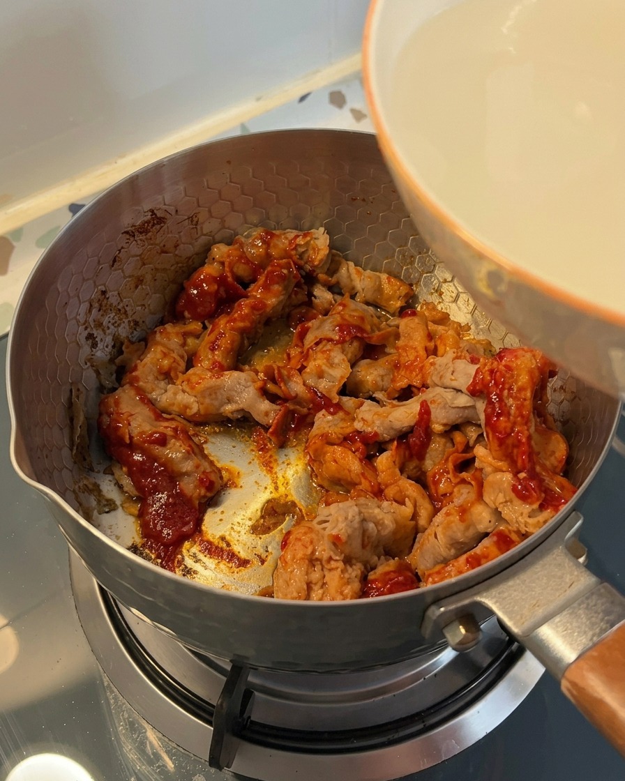 Trübes Reiswaschwasser wird aus einer weißen Schüssel in einen Topf mit dem angebratenen Rindfleisch gegossen.