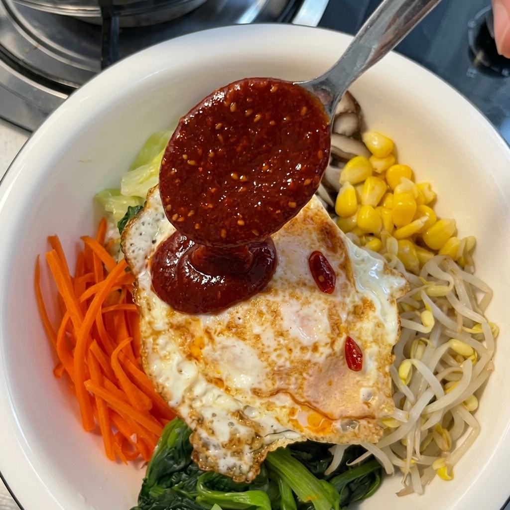 Eine dickflüssige rote Chilipaste-Sauce wird mit einem Metalllöffel über ein Spiegelei gegossen, das auf einer bunten Auswahl an Bibimbap-Gemüse liegt.