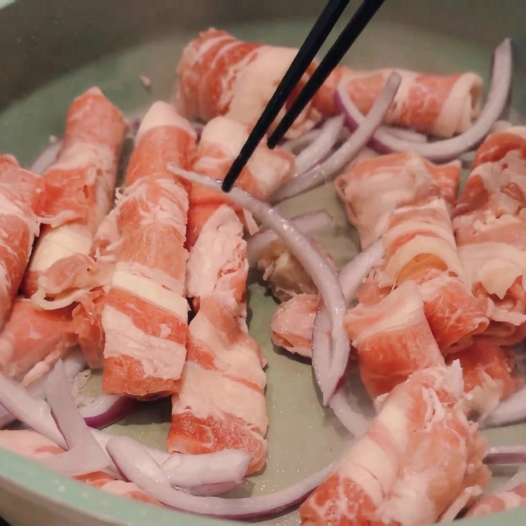 Essstäbchen rühren dünne Scheiben von rohem Rindfleisch und roten Zwiebeln in einer hellgrünen Pfanne.