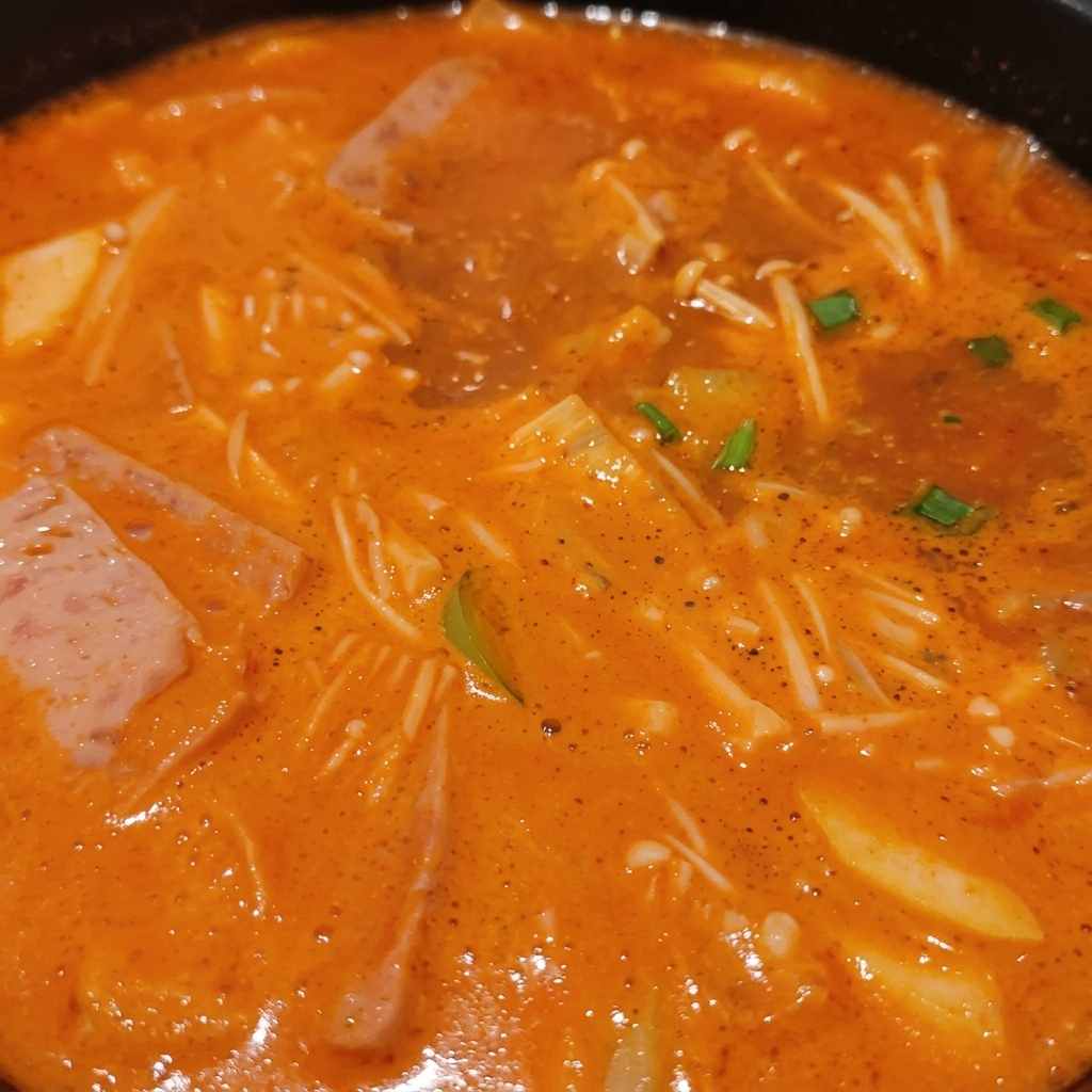 Ein brodelnder koreanischer Eintopf voller Enoki-Pilze, Fleischscheiben, grüner Frühlingszwiebeln und Tofu in einer reichhaltigen orangefarbenen Brühe.