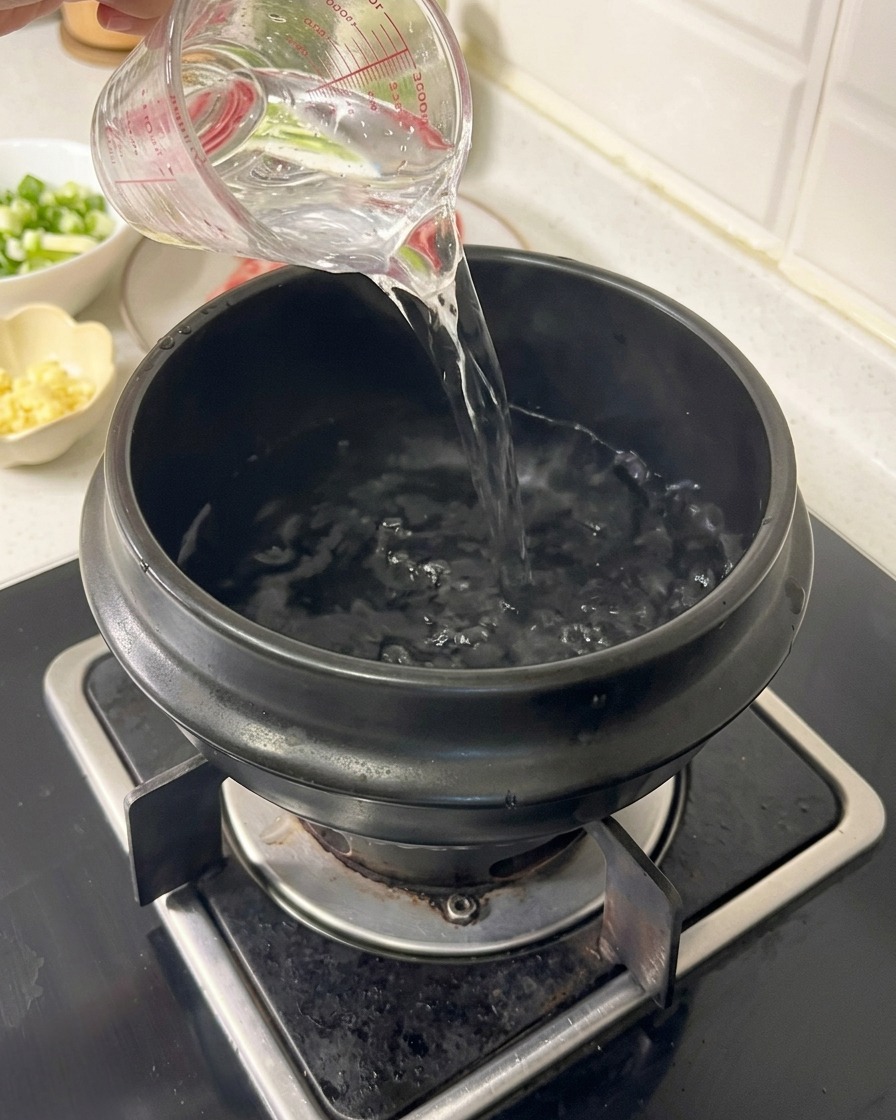 Wasser wird aus einem klaren Messbecher in einen schwarzen irdenen Tontopf auf einem Gasherd gegossen.