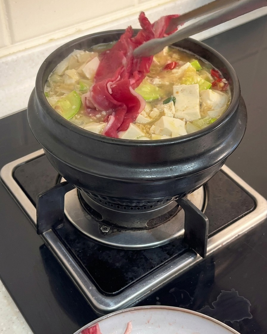 Eine Küchenzange legt dünne Scheiben rohes Rinderbrustfleisch in einen kochenden koreanischen Eintopf mit Tofu und Zucchini.