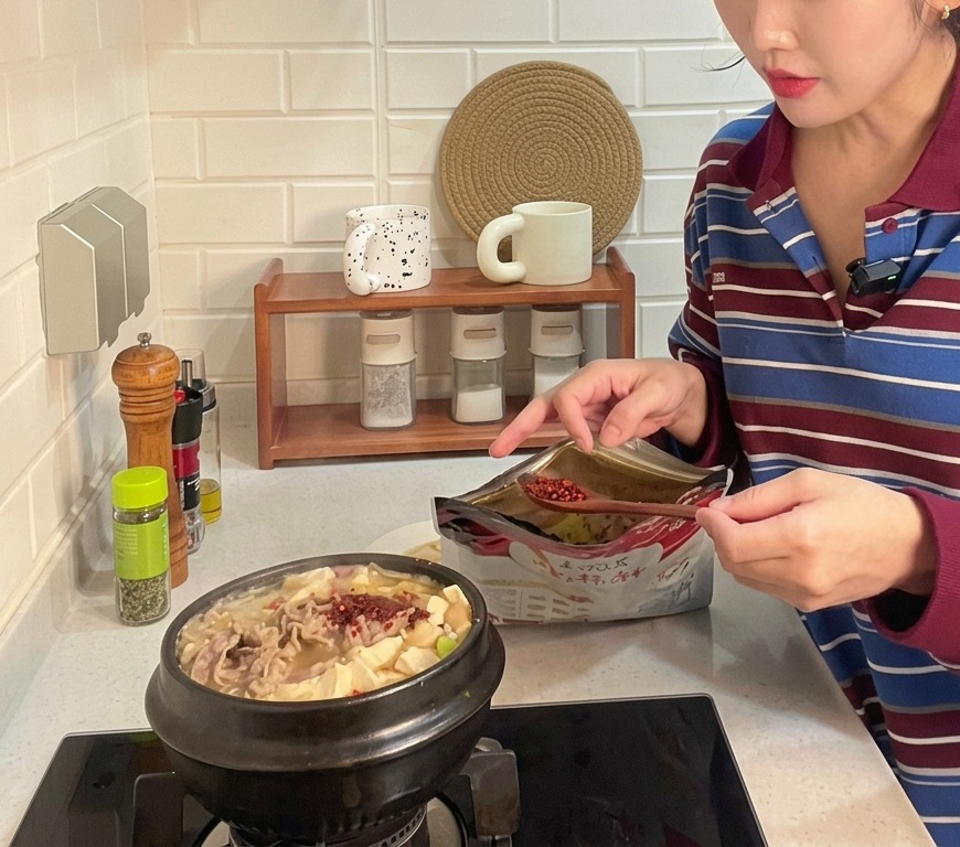 Ein Löffel grobes rotes Chilipulver wird in einen brodelnden Tontopf mit koreanischem Sojabohnenpasten-Eintopf gegeben, der mit Tofu und Rindfleisch gefüllt ist.