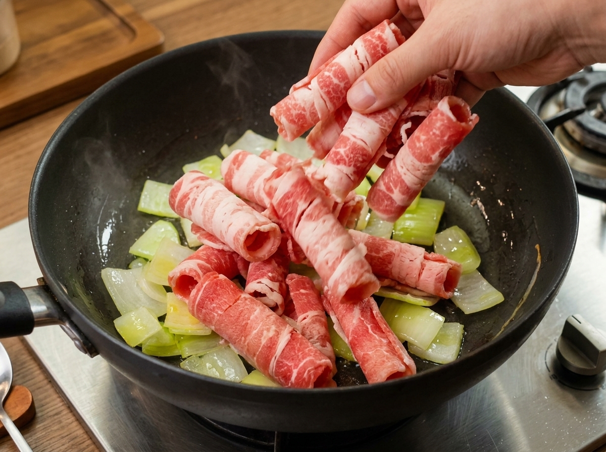 Dünne Rindfleischscheiben werden zu glasig angedünsteten Zwiebeln in eine Pfanne gegeben.