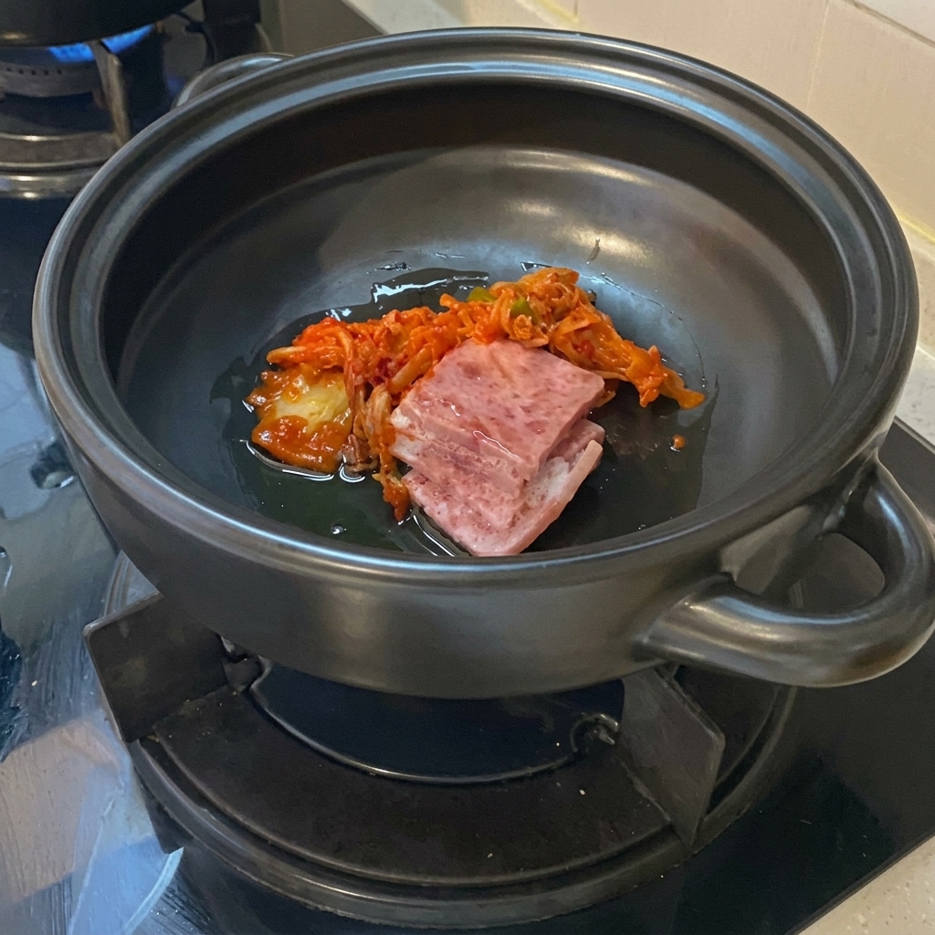 Rohe Schweinebauchscheiben und farbenfrohes gehacktes Kimchi brutzeln im Öl auf dem Boden eines dunklen Keramiktopfs.