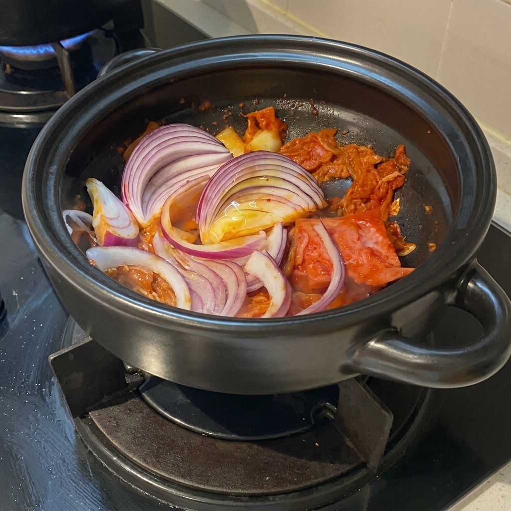 Dick geschnittene rote Zwiebelstreifen liegen auf dem angebratenen Kimchi und Schweinebauch im Tontopf.