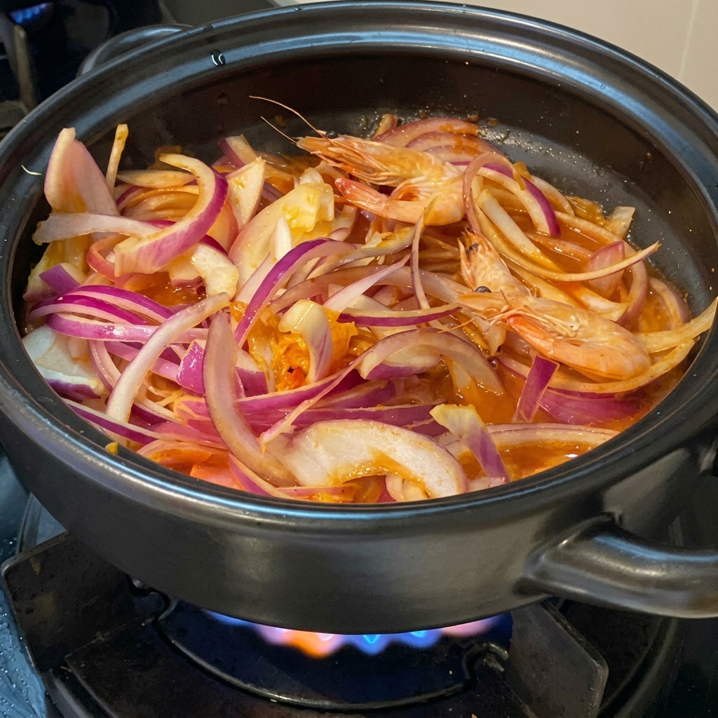 Frische, ganze rosa Garnelen liegen eingebettet zwischen roten Zwiebelscheiben und Kimchi in einem schwarzen Tontopf auf dem Herd.