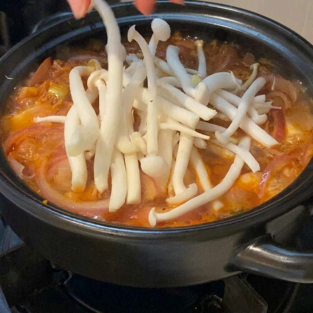 Frische weiße Buchenpilze werden sorgfältig von Hand auf die kochende rote Kimchi-Suppe im Tontopf gelegt.