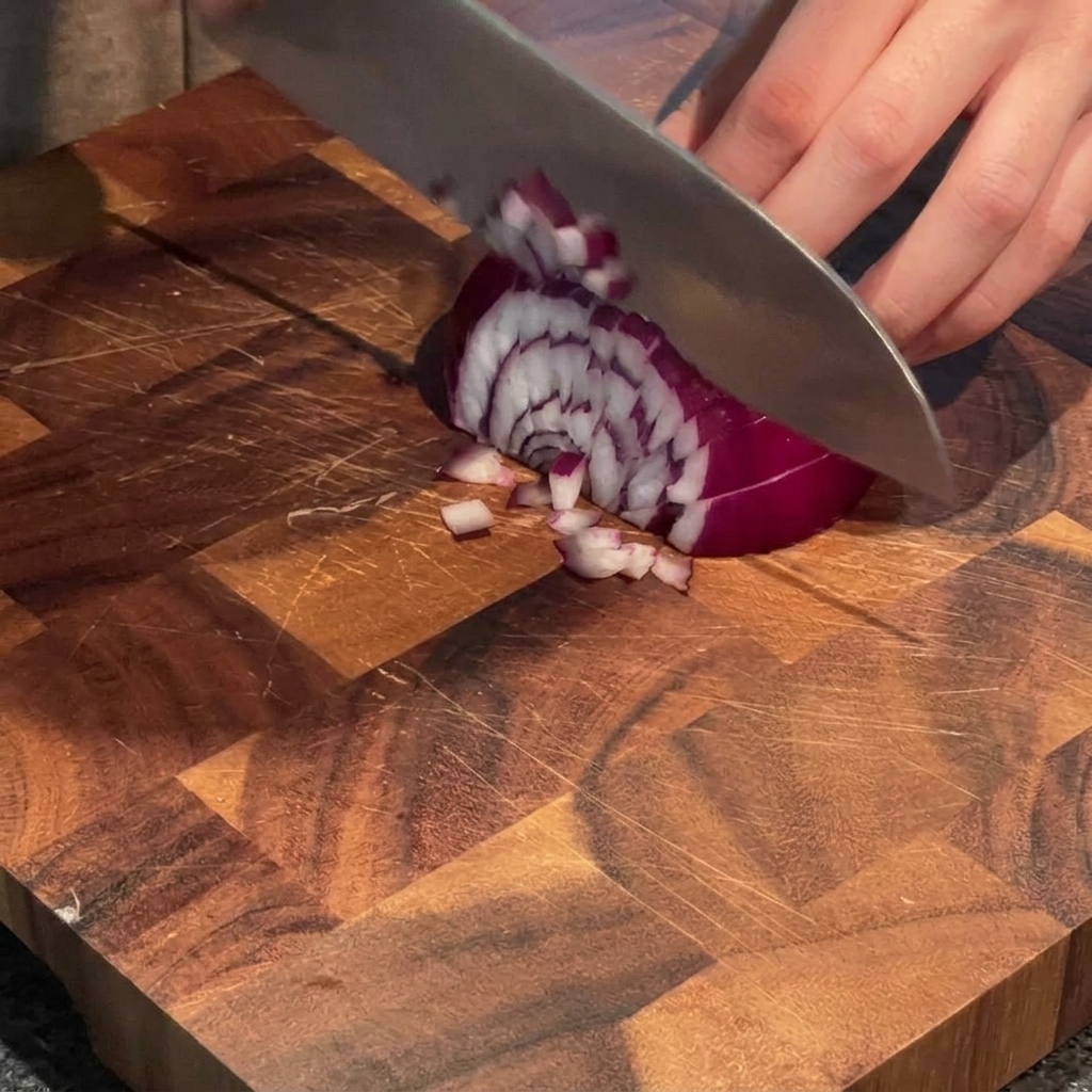 Eine Hand, die mit einem Kochmesser eine rote Zwiebel auf einem Holzschneidebrett in kleine Stücke würfelt.