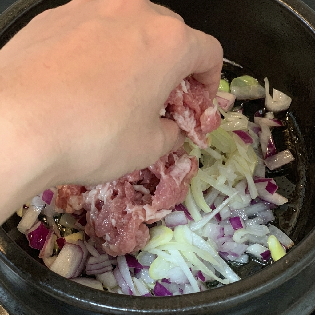 Rohe Schweinebauchstücke werden in einen Kochtopf mit gewürfelten roten Zwiebeln und Frühlingszwiebelstreifen gegeben.