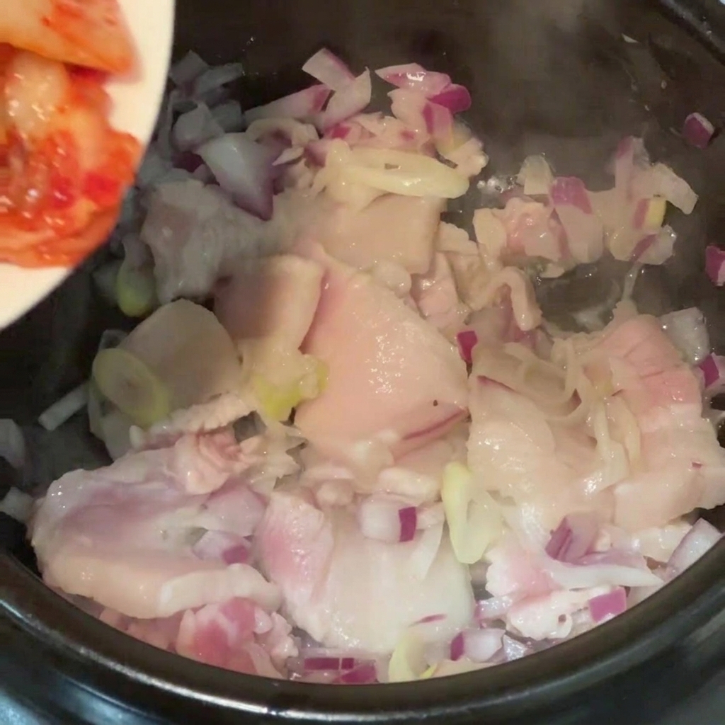 Geschnittener Kimchi-Kohl wird von einem Teller in einen Kochtopf mit Schweinebauchstücken und roten Zwiebeln gegeben.
