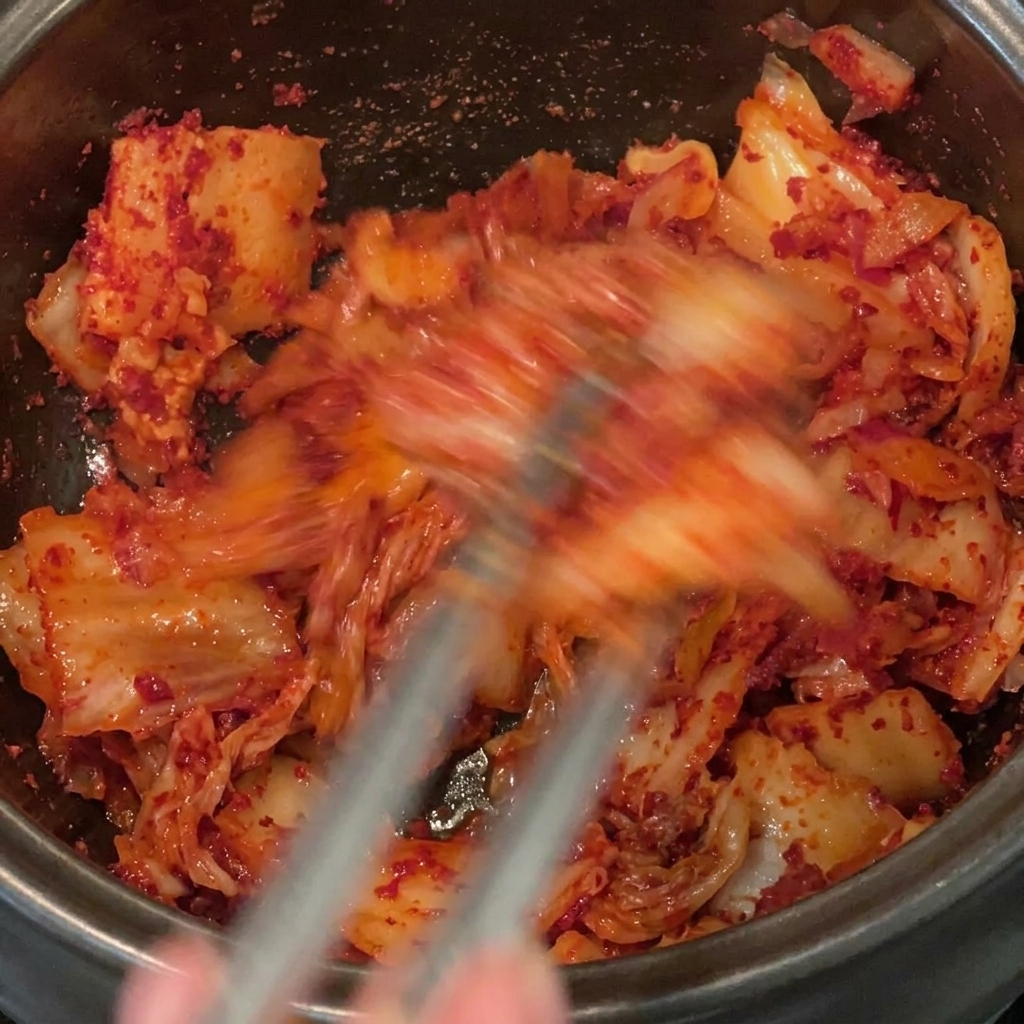 Rotes Chilipulver wird gründlich mit geschnittenem Kimchi und Schweinebauch in einem schwarzen Topf verrührt.