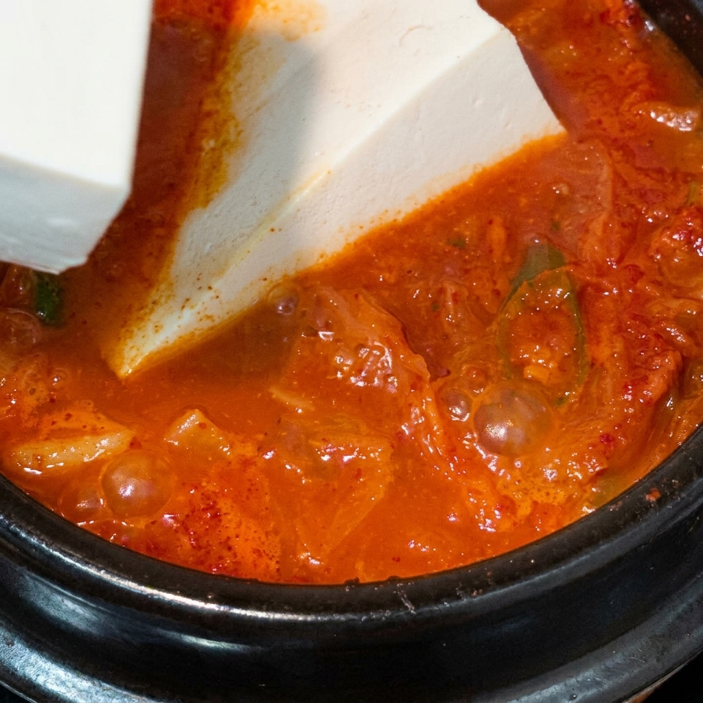 Ein großer Block weißer Seidentofu wird vorsichtig in einen kochenden Topf mit roter Kimchi-Suppe gelegt.