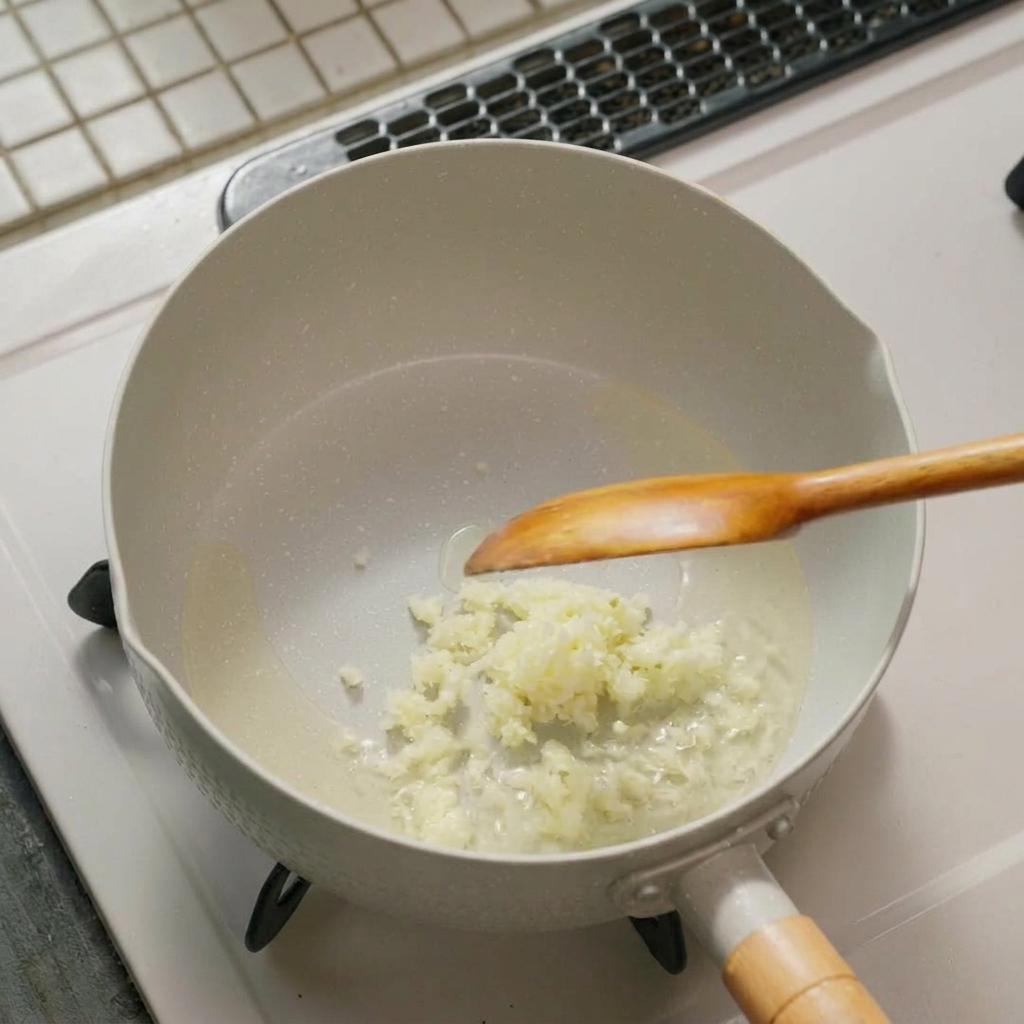 Fein gehackter Knoblauch und Frühlingszwiebeln brodeln in Öl auf dem Boden einer weißen Pfanne, in der ein Kochlöffel liegt.