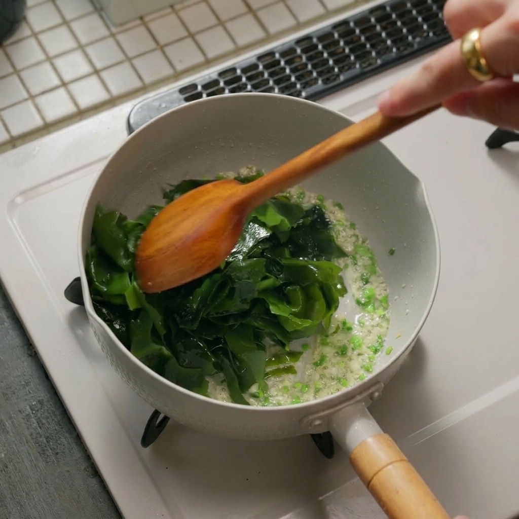 Ein Haufen leuchtend grüner, eingeweichter Algen wird zu den angeschwitzten Aromaten in eine weiße Pfanne gegeben und mit einem Holzwender umgerührt.