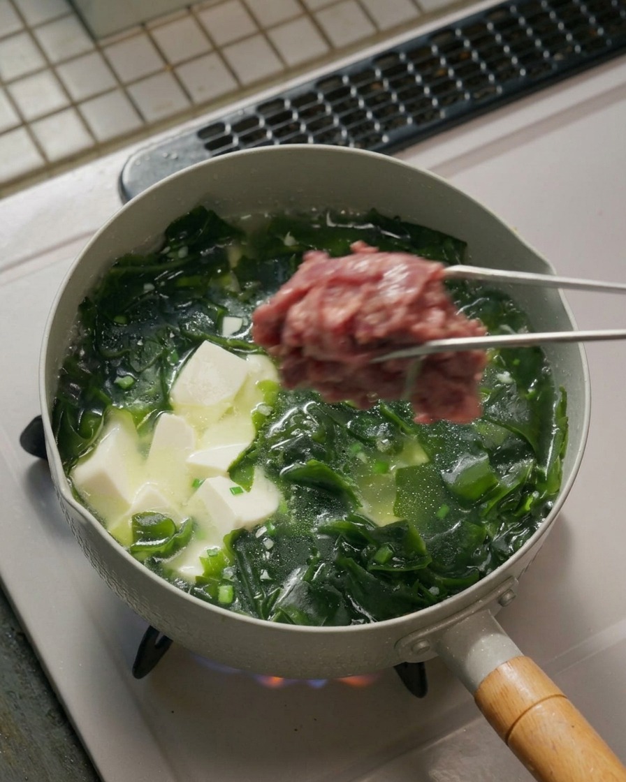 Eine Metallzange hält eine Portion rohe, marinierte Rindfleischscheiben über einem kochenden Topf voller Seealgen- und Tofusuppe.