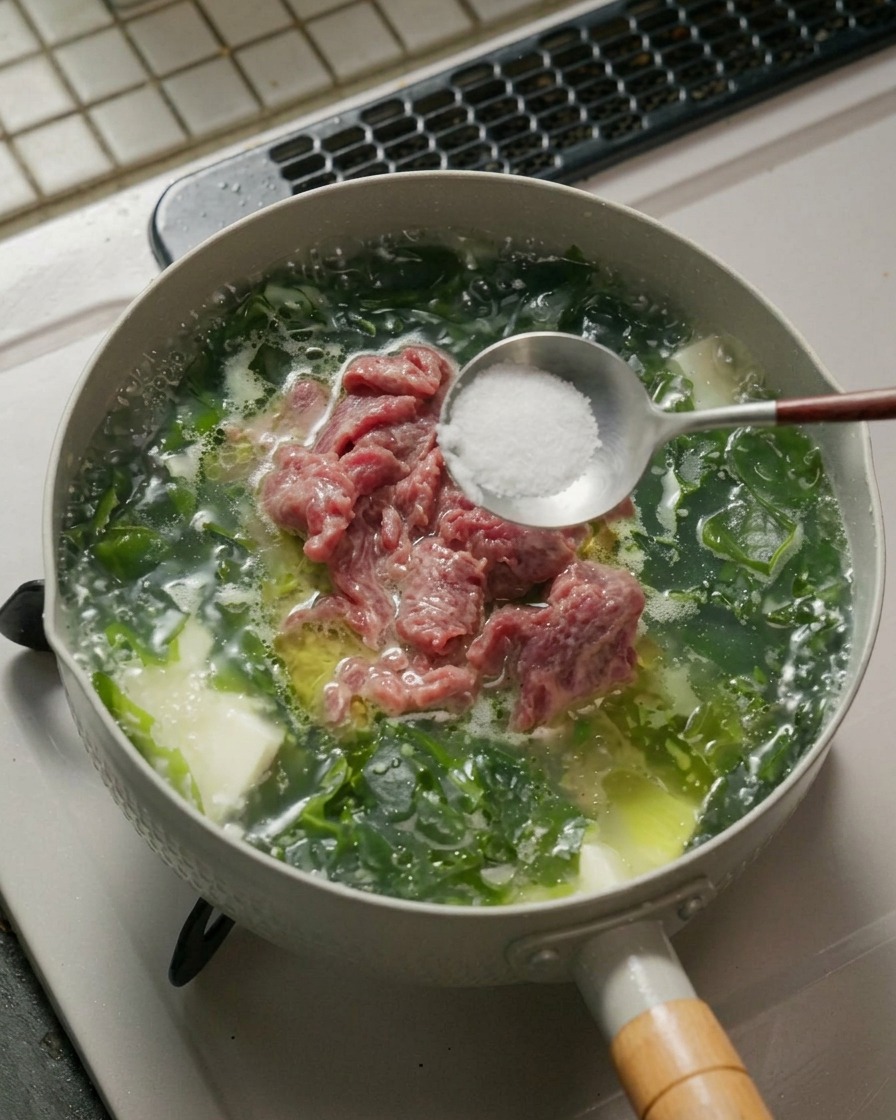 Ein Metalllöffel hält weißes Salz und Pfefferpulver über einen Topf mit köchelnder Seealgen-Rindfleisch-Tofu-Suppe.