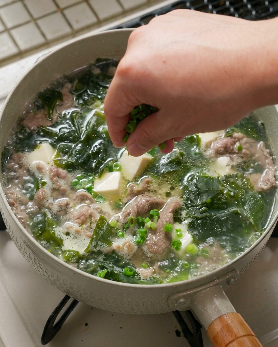Eine Hand streut eine großzügige Prise fein gehackter Frühlingszwiebeln über eine heiße, köchelnde Pfanne mit Seealgensuppe, weißen Tofuwürfeln und gegartem Rindfleisch.