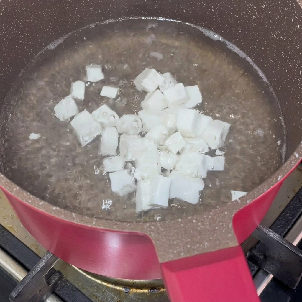 Kleine Würfel aus weichem, weißem Tofu, die in kochendem Wasser in einem rosafarbenen, beschichteten Kochtopf blanchiert werden.