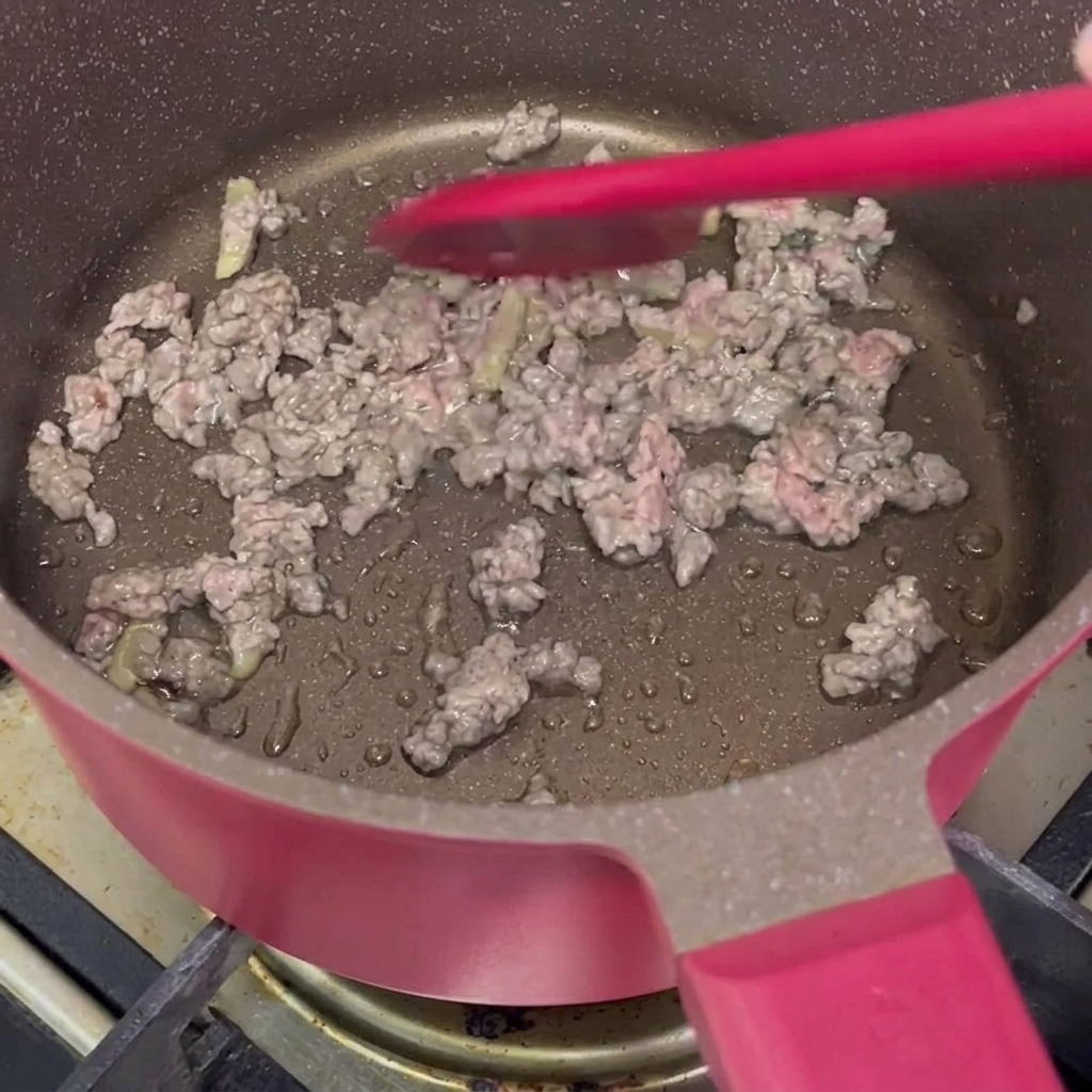 Mariniertes Rinderhackfleisch, das mit einem roten Silikonspatel in einem rosafarbenen Topf angebraten wird, bis das Fleisch die Farbe ändert.