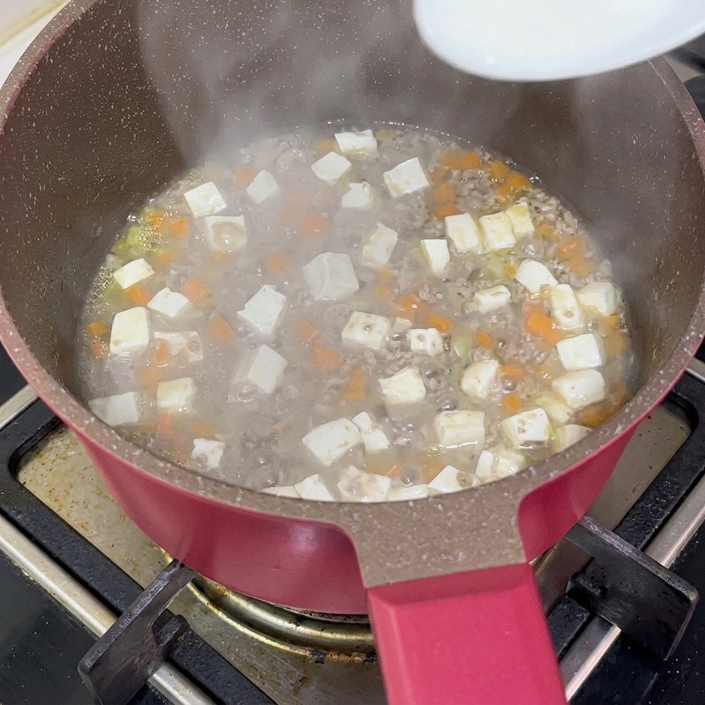 Ein weißer Löffel gießt eine leicht milchige Stärkemischung in den kochenden Eintopf aus Fleisch, Tofu und Karotten, um die Soße zu binden.