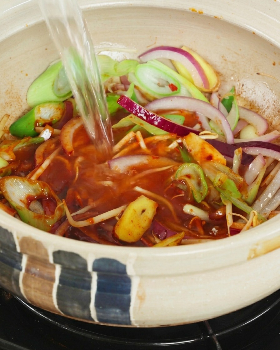 Ein Strahl klares, kochendes Wasser wird in einen Keramichtopf mit Zwiebeln, Frühlingszwiebeln und roter Chilibrühe gegossen.