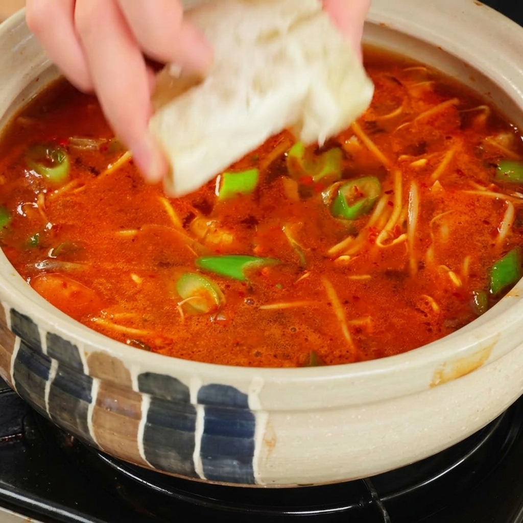 Eine Hand gibt einen quadratischen, hellen Brühwürfel in einen kochenden Topf mit leuchtend roter, scharfer Suppe.