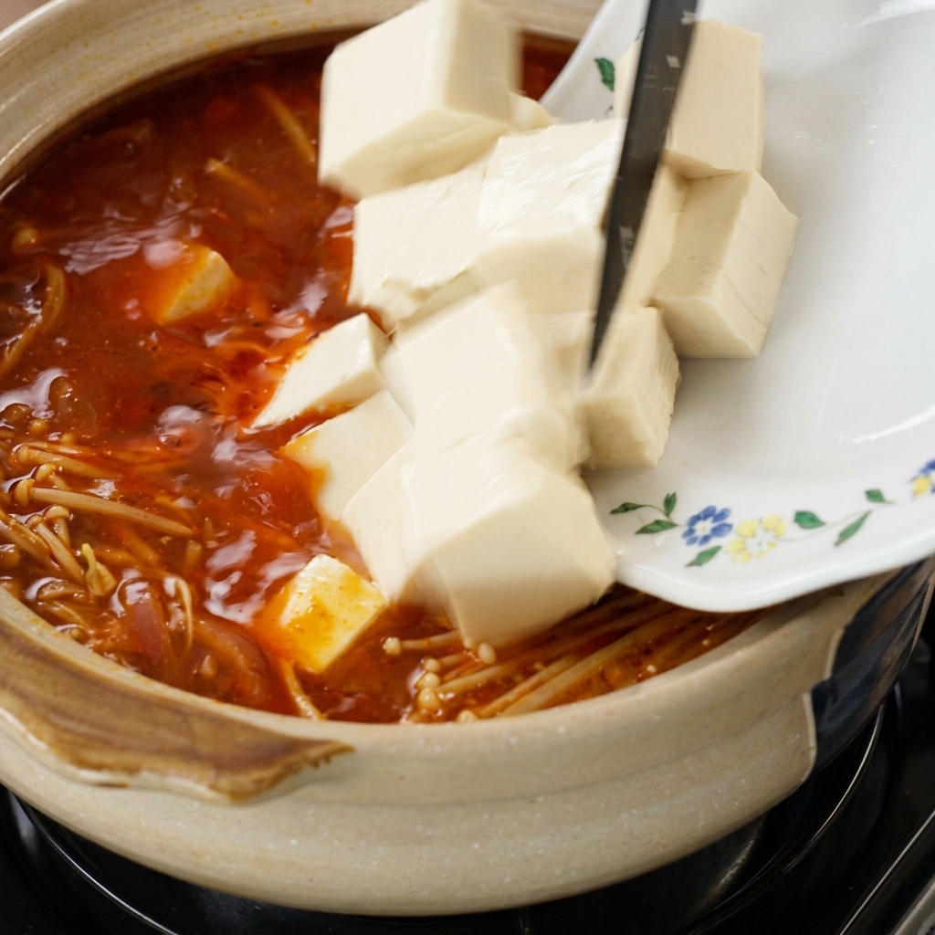 Frische weiße Tofuwürfel werden von einem gemusterten Teller in die kochende rote Suppe mit Enoki-Pilzen geschoben.