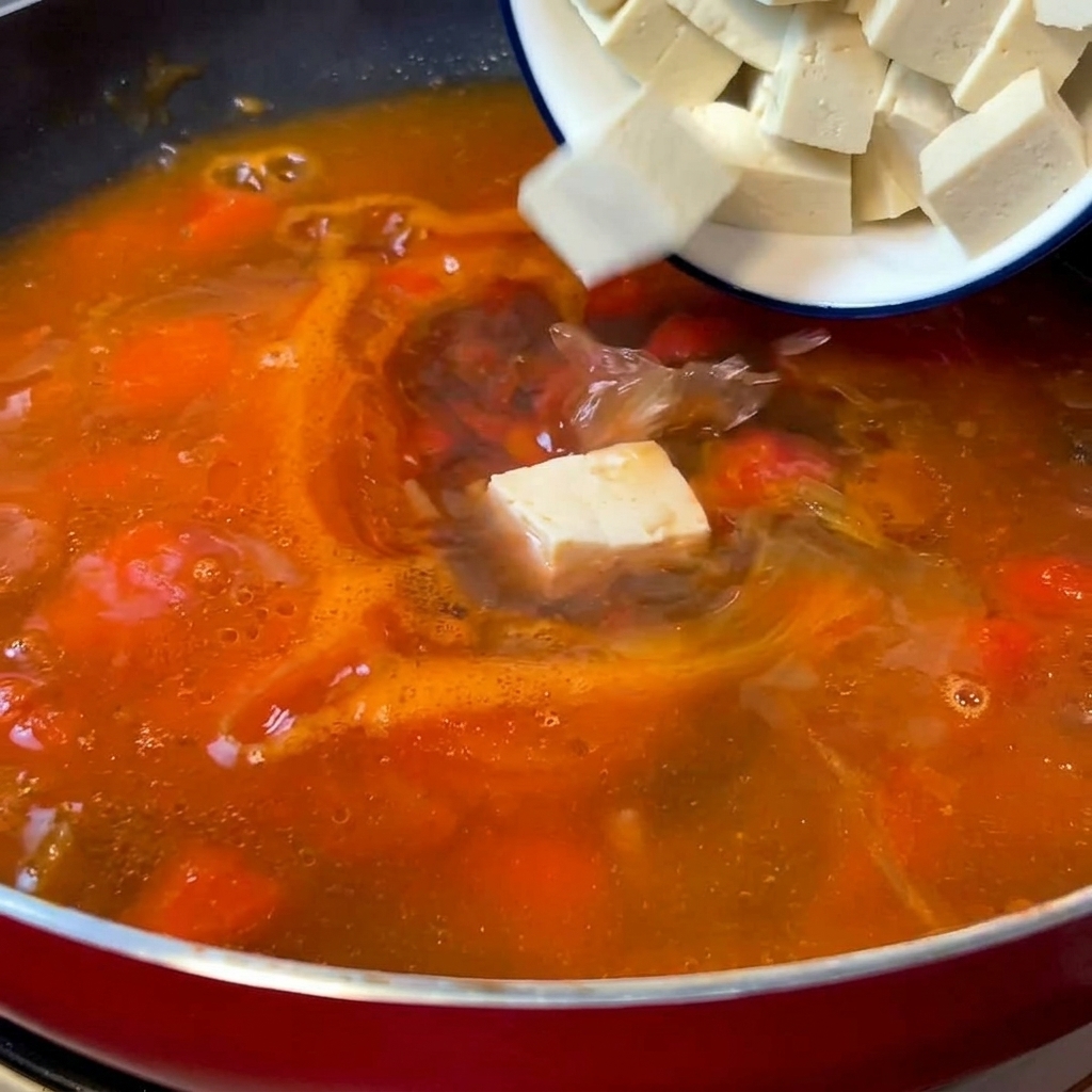 Weiße Seidentofuwürfel werden vorsichtig aus einer kleinen Schüssel in eine rote Pfanne mit kochender, orange-roter Tomatensuppe gegeben.