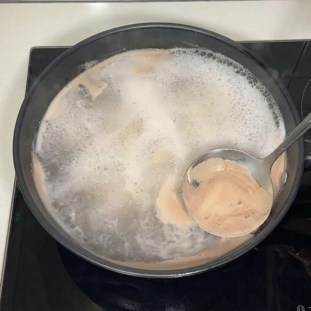 Eine Schöpfkelle aus Metall schöpft braunen Schaum und Trübstoffe von der Oberfläche einer kochenden Brühe in einer schwarzen Pfanne.