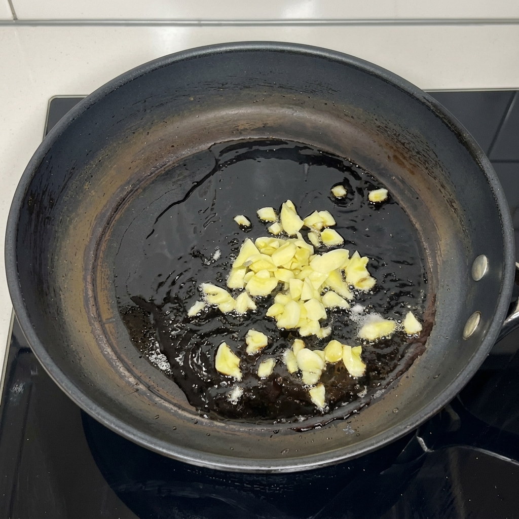 Fein gehackte Knoblauchzeilen brodeln in heißem Pflanzenöl am Boden einer dunklen Pfanne.