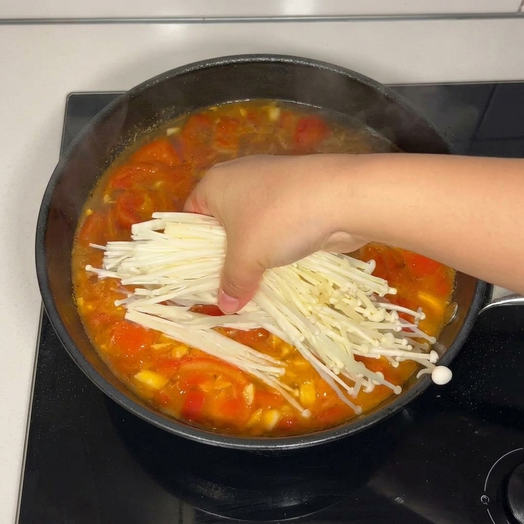 Eine Hand gibt ein Büschel frischer, weißer Enoki-Pilze in die köchelnde Tomatensuppe in der Pfanne.