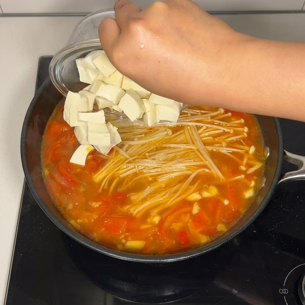 Gewürfelter, weicher weißer Tofu wird aus einer klaren Glasschale in die kochende Tomatensuppe mit Enoki-Pilzen gleiten gelassen.