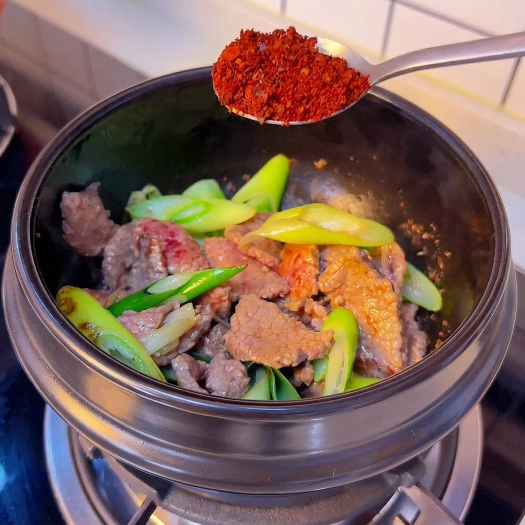 Ein silberner Löffel mit grobem rotem Chilipulver über einem schwarzen Topf mit angebratenem Rindfleisch und Frühlingszwiebeln.