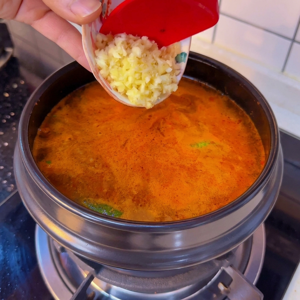 Gehackter Knoblauch wird aus einer kleinen Schüssel in einen blubbernden Topf mit scharfem koreanischen Seidentofu-Eintopf geschabt.