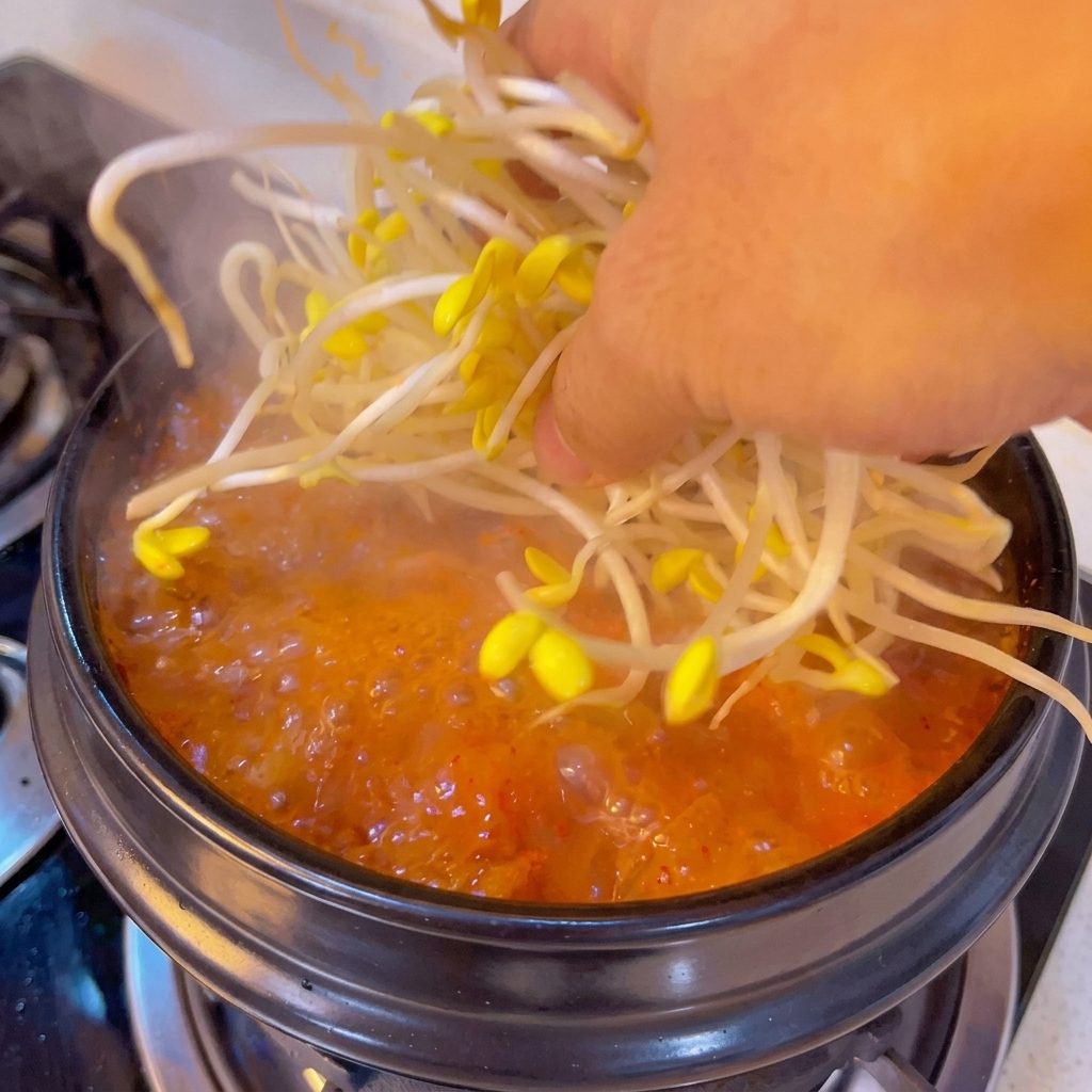 Eine Hand gibt eine Handvoll frischer gelber Sojasprossen in einen blubbernden, orange-roten Suppentopf.
