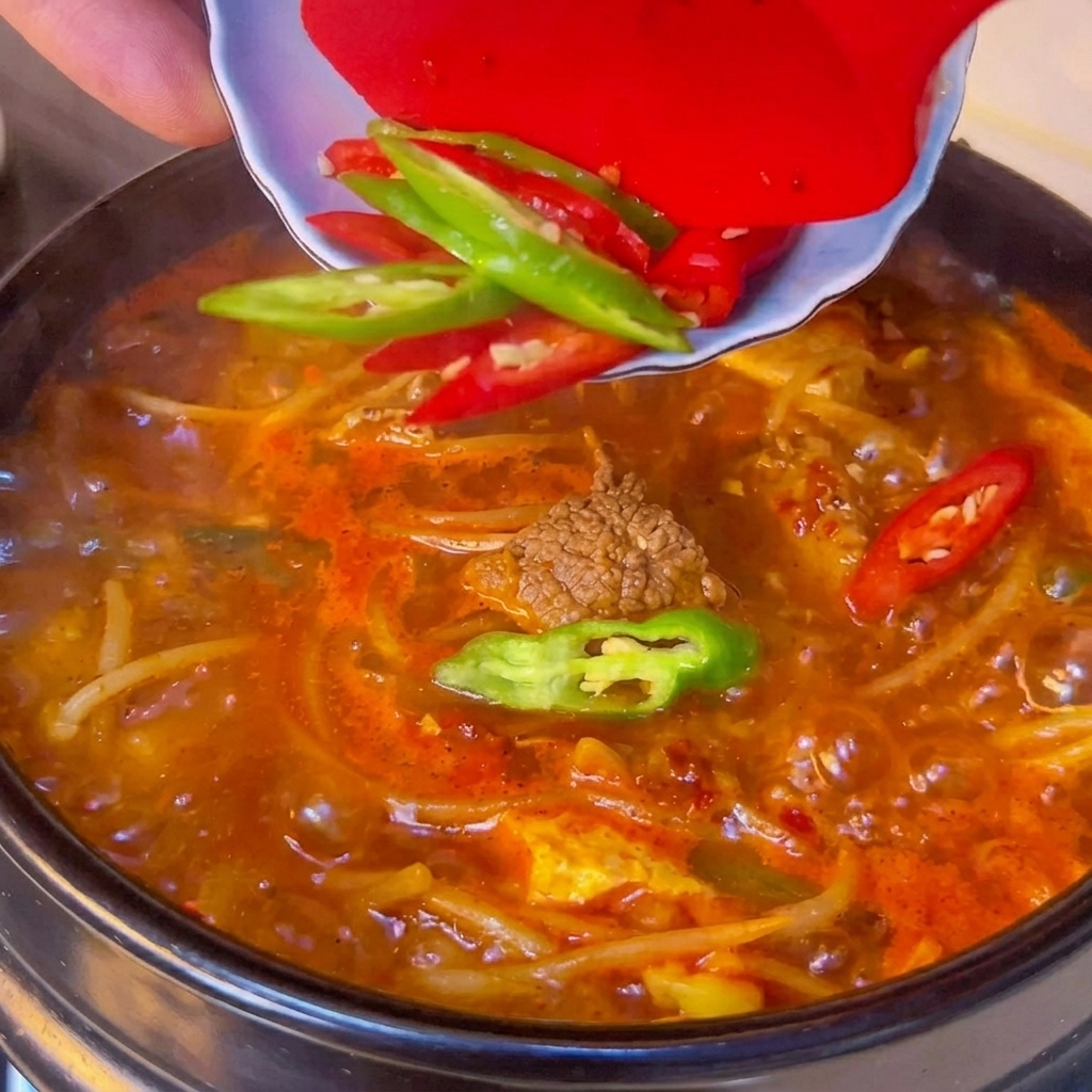 Frische grüne und rote Chilischeiben werden aus einer kleinen weißen Schüssel auf die Oberfläche eines kochenden scharfen Tofu-Eintopfs geschoben.