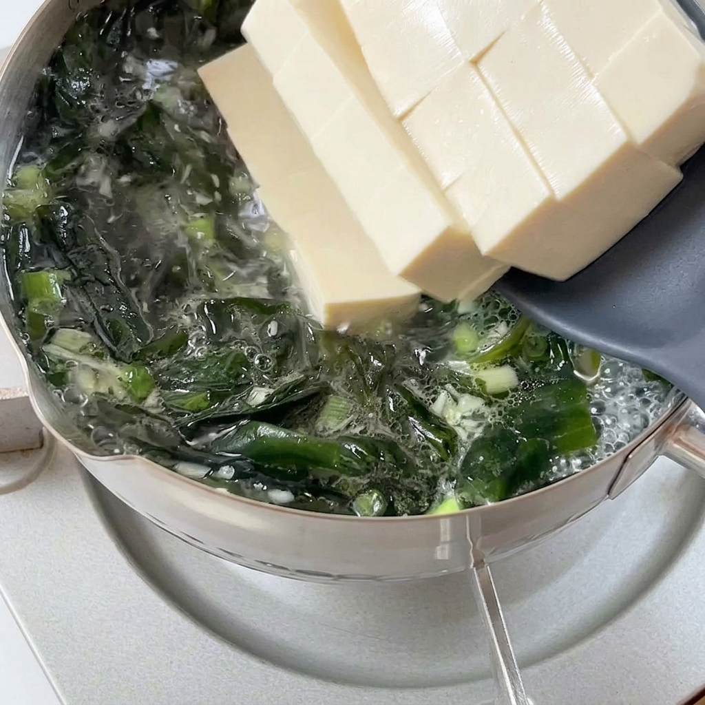 Dicke Stücke von weichem, weißem Tofu gleiten von einem grauen Spatel in einen sprudelnden Topf mit Wakame-Brühe.