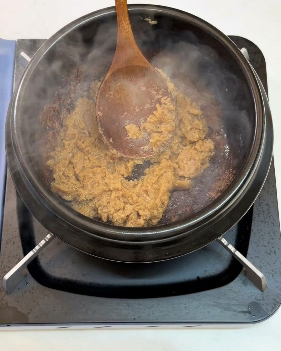 Ein Holzlöffel rührt gehackten Knoblauch und Sojabohnenpaste in einem heißen Steintopf um.